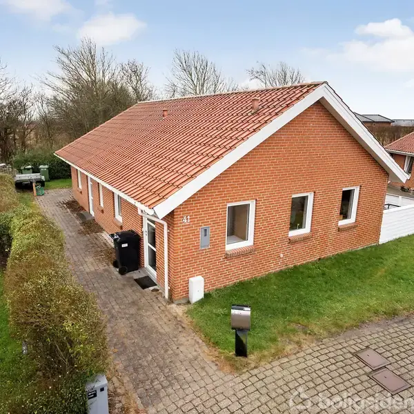 En rød murstensvilla med rødt tegltag står i et grønt haveområde. Den ligger ved en belagt sti, omgivet af buske, træer og en græsplæne. Ingen tekst til stede.