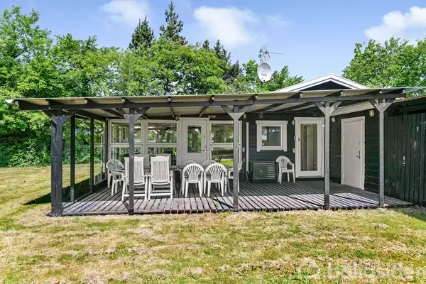 En træterrasse med hvide plaststole og bord under overdække, beliggende foran et mørkt træhus. Omgivet af en grøn have med græs og træer i baggrunden.