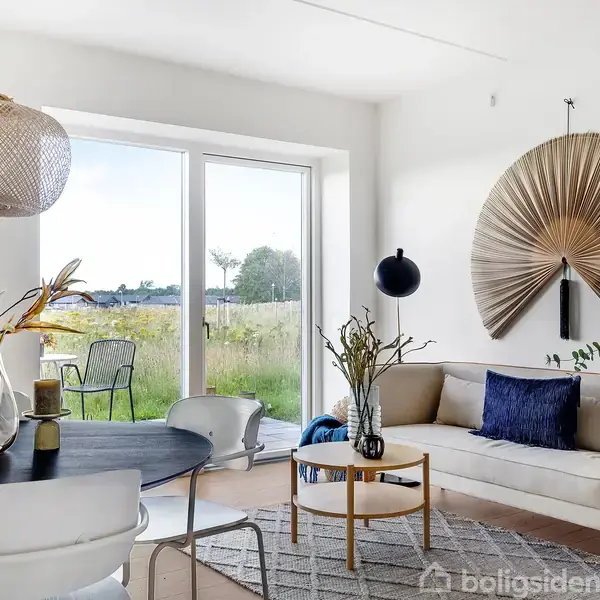 Stue med rund spisebord og stole, dekoreret med planter i vase. Sofa med blå pude står ved store vinduer, der viser udsigt til grønt landskab. Elegant vægdekoration tilføjer stil.
