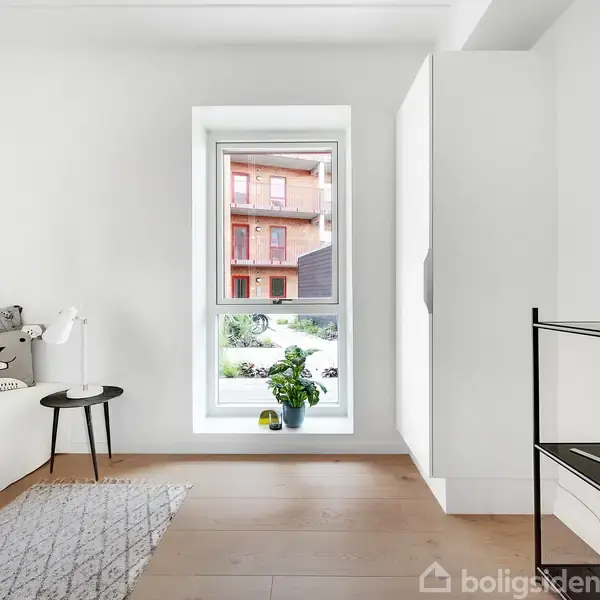 Et lyst rum har en sofa med puder og tæppe til venstre, en reol med planter til højre og en stor vindue, der viser en udsigt til en have og bygninger udenfor.
