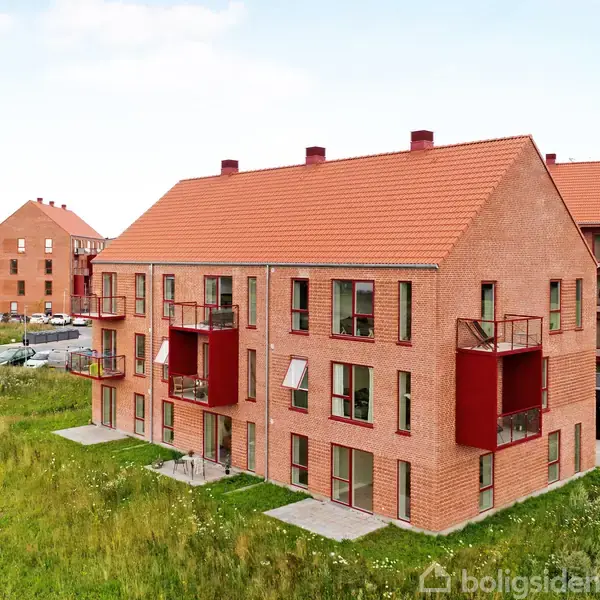 En moderne murstensbygning med røde altaner står i forgrunden. Græs og parkering omgiver bygningen. Flere lignende bygninger ses i baggrunden.