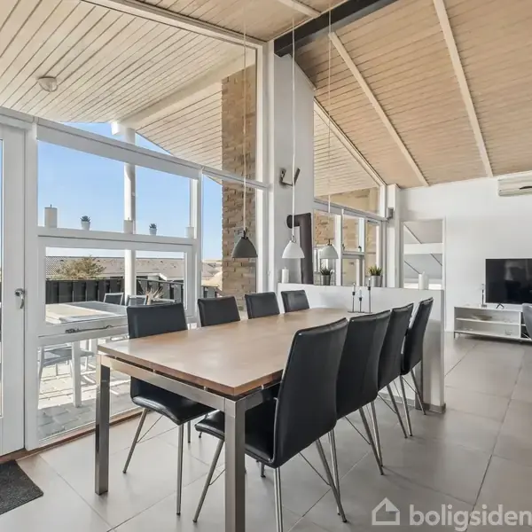 Hus i mursten står stille i et klitlandskab med høj bevoksning. Klar blå himmel danner en lys baggrund. Terrasse med stole til højre.