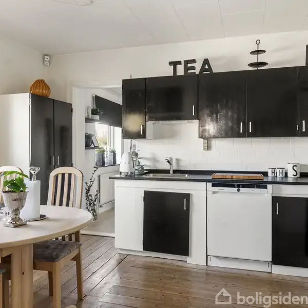 Et køkken med sort-hvide skabe og bordplade, en rund spisebord med stole i forgrunden. Tekst på væggen: "TEA" og "KAFFE". Lyst trægulv dominerer rummet.