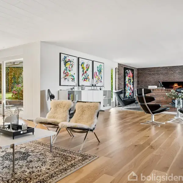 En moderne stue med en stor grå hjørnesofa står ved et vindue, der giver udsigt til et hus udenfor. En murstensvæg indeholder pejs, TV og reoler. Sorte flisegulve.