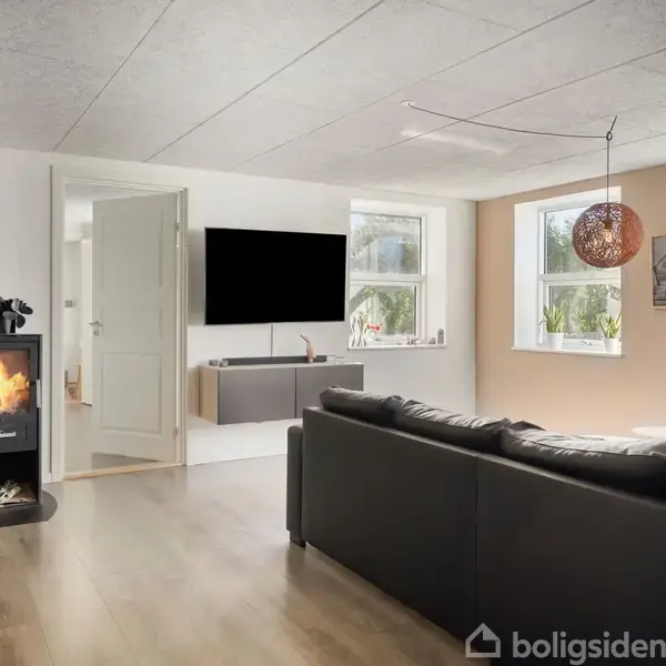 En sort sofa står i en stue med trægulv, hvor en brændeovn er tændt. Baggrunden har hvide vægge, en stor fladskærm, og vinduer med udsigt til grønne områder.