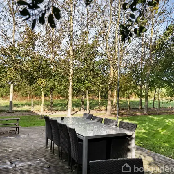 Terrassebord omgivet af stole står på en træterrasse. Skoven ses i baggrunden i en solrig have med græsplæne.