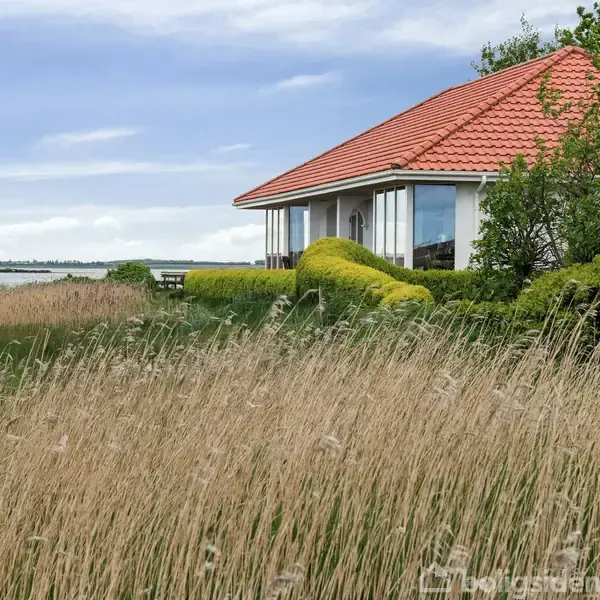Et hus med rødt tegltag står stille ved siden af tætbevokset græs og buske. I baggrunden ses havet og en åben himmel.