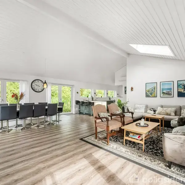 En rummelig stue med lyse trægulve, hvide vægge og loft. Et langt spisebord med sorte stole og en stor vægur dominerer venstre side, mens en sofagruppe med tæppe er på højre side. Store vinduer giver udsigt til grønne omgivelser.