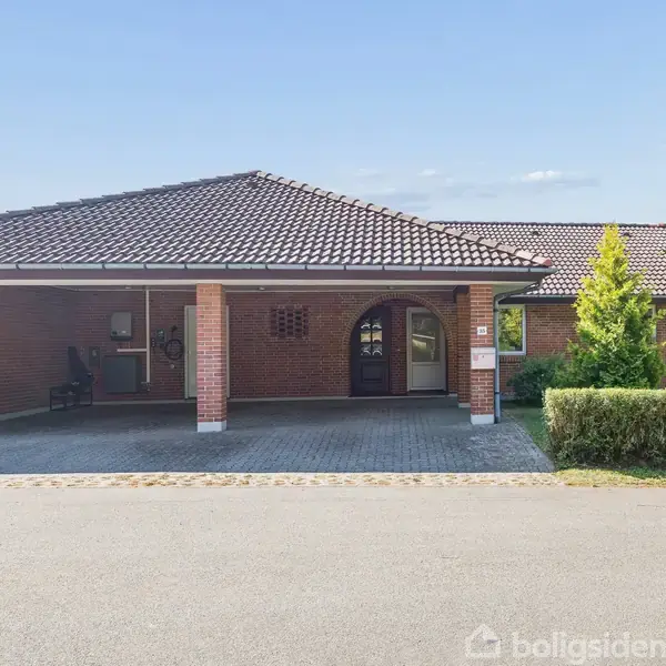 Hus i røde mursten med klinketag står stille langs vejen. Grønt buskads og et lille træ danner en havekant til højre. Carport er del af husets design.