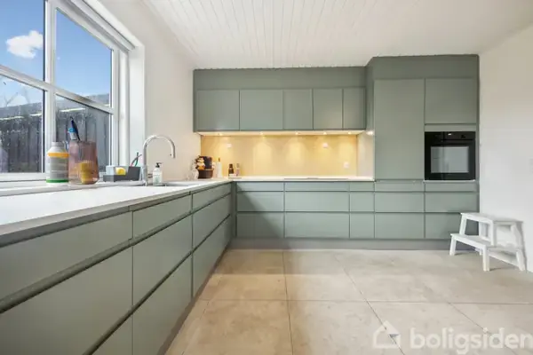 Køkken i moderne stil, hvor grønne skabe fylder væggene, en indbygget ovn er til højre, og store vinduer giver lyse omgivelser. Skammel ved siden af ovnen.