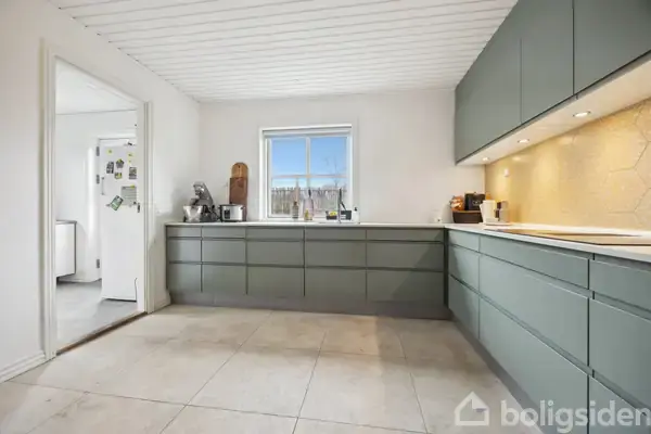 Et stilrent køkken med grå skuffer og hvide vægge. Et vindue centralt over arbejdsfladen. Et andet rum ses gennem en åben dør. Belysning under overskabe oplyser frugter.