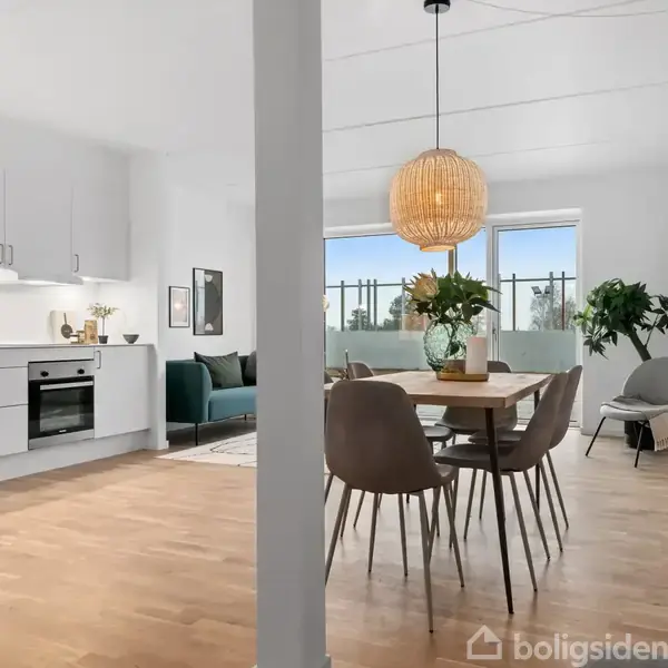 Køkken-alrum med hvide skabe og integreret ovn. Spisebord med seks stole under en lampe. Sofa i stuen ved vindue med planter. Lyse trægulve og minimalistisk indretning.