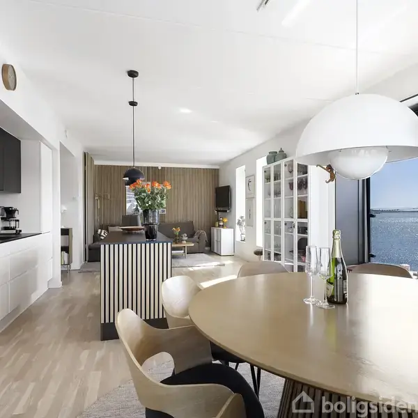 Rundt spisebord med stole står i moderne køkken-alrum, hvor en lysekrone hænger over bordet. Store vinduer giver udsigt til vandet. En vase med blomster og flasker står på bordet.