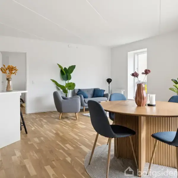 Rundt træbord med fire blå stole står i et lyst, moderne stue-køkken-alrum. Sofa og lænestol med plante og lampe i baggrunden, ved siden af køkkenø med blomster. Ingen tekst.