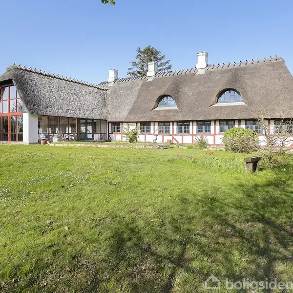 Stråtækt bindingsværkshus ligger på en stor grøn græsplæne med små buske og træer omkring. Huset har flere vinduer og en glasudbygning. Klar blå himmel i baggrunden.