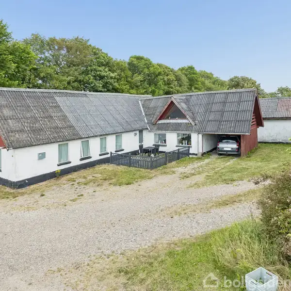 Et langt bondehus med hvid facade og rødt tag står stille. Grusbelagt indkørsel fører til en bilport. Grønne træer omgiver ejendommen i baggrunden.