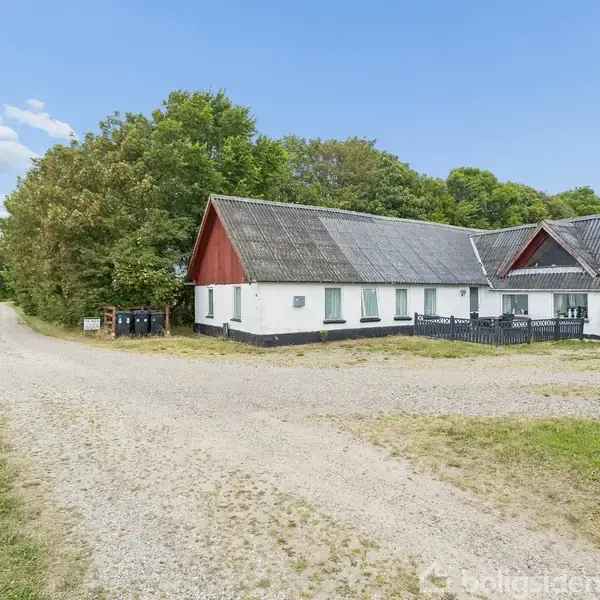 Landhus med røde gavle står stille ved en grusvej. Til venstre ses åbent marklandskab, mens skov grænser op mod husets baggrund. En bil er parkeret under et halvtag.