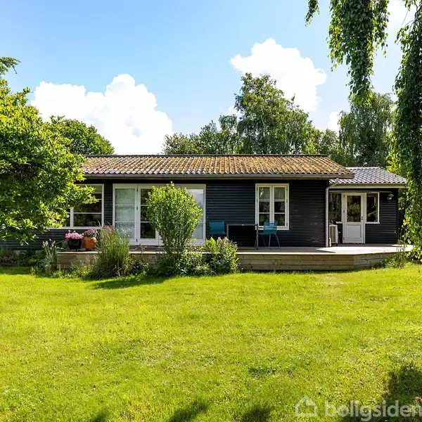 Et sortmalet hus står stille i en frodig have med grøn græsplæne og buske omkring. Trædækket foran huset har havemøbler. Himlen er blå med hvide skyer.