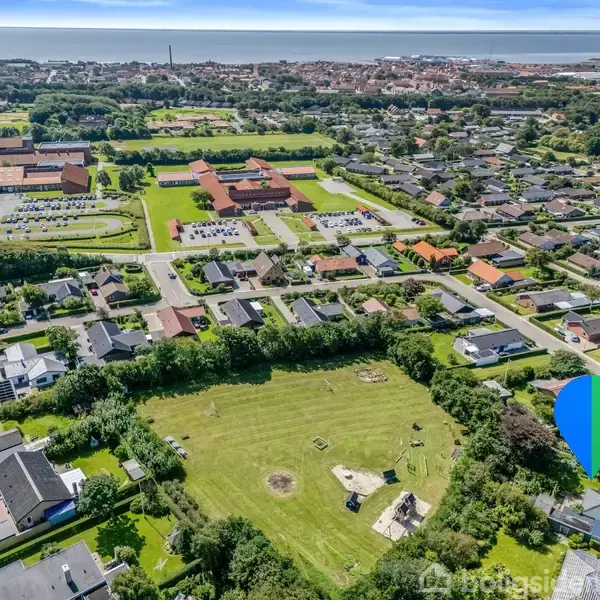 En boligkvarter med huse omgivet af grønne områder. Børn leger på en legeplads i en stor græsmark. I baggrunden ses skolebygninger og en by med udsigt til vandet.
