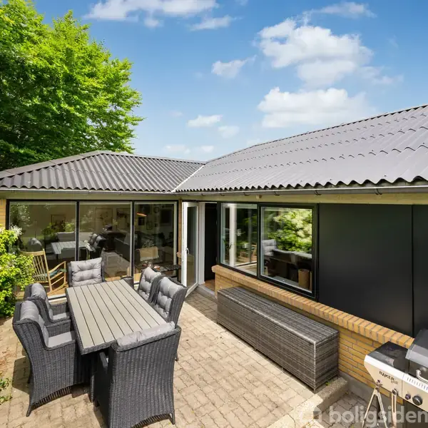 Terrasse med stort spisebord og stole, omgivet af grønne buske. Huset har store vinduer mod terrassen og grå tagsten. Grill står til højre. Skyfri blå himmel i baggrunden.