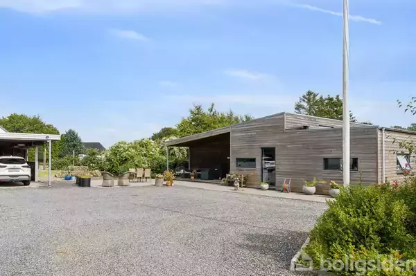 Et moderne træhus med fladt tag står stille, omringet af planter og en grusbelagt gårdsplads. En bil er parkeret under en carport til venstre.