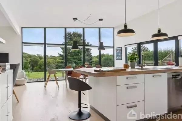 Fladt, modernistisk hus af træ placeret på en grusbelagt indkørsel. Flagstang står ved siden af, omkranset af træer og buske i et grønt haveområde.