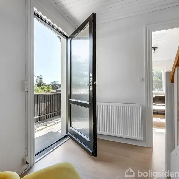 En glasdør står åben og fører ud til en balkon. Indenfor ses en gul lænestol og en radiator ved en væg i en lys stue med trægulv.