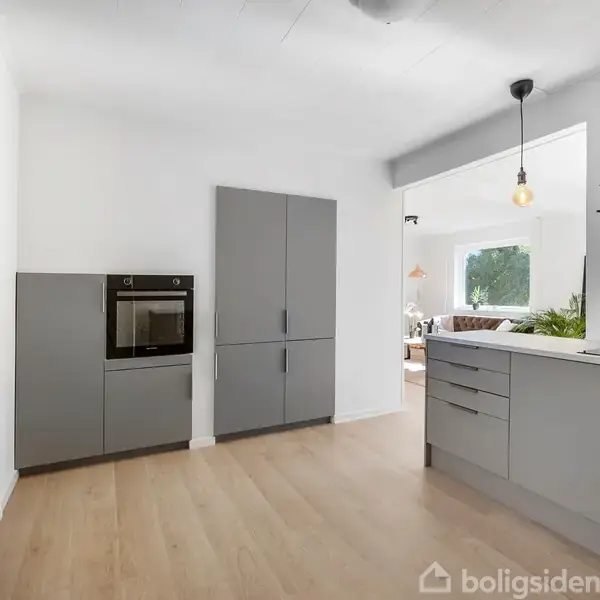 Et moderne køkken med grå skabe og indbygget ovn, åbner ud mod en stue med store vinduer, trælaminatgulv og hængende lampe.