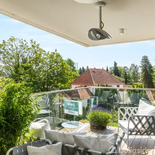 En balkon med komfortable stole og puder vender mod en soloplyst udsigt over hustage og træer i et beboelsesområde. En plante tilføjer grønne nuancer til omgivelserne.