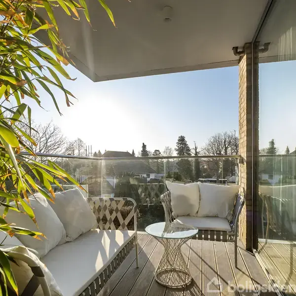 En moderne altan indeholder hvide polstrede møbler og et lille rundt bord. Omkring altanen ses glasgelænder og tæt beplantning. I baggrunden er der udsigt til træer og hustage.