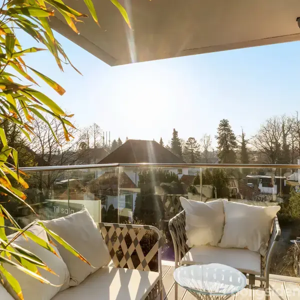 Balkon med hvide puder på stole, sollys stråler ind. I baggrunden ses træer og hustage.