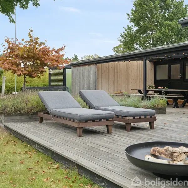 To solsenge står på en træterrasse ved siden af en lavendelbusk og en ildskål med brænde; i baggrunden findes et hus med overdækket udendørs spiseområde.