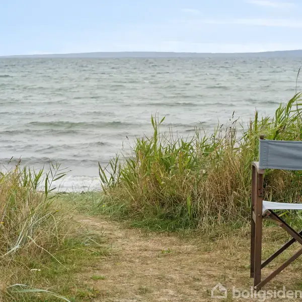 En foldestol står stille ved en sti foran havet. Den er omgivet af højt græs, med en klar udsigt over det bølgende vand og en fjern horisont.