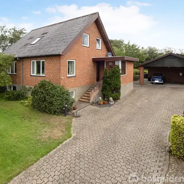En rødstensvilla med det skråtag står stille ved siden af en carport med en bil. Omgivet af en velplejet have med buske og græsplæne.