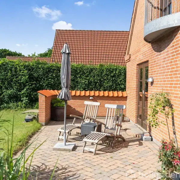 To liggestole og en parasol står på en flisebelagt terrasse foran en rød murstensbygning. Terrassebordet er omgivet af en græsplæne og hække under en blå, skyfri himmel.