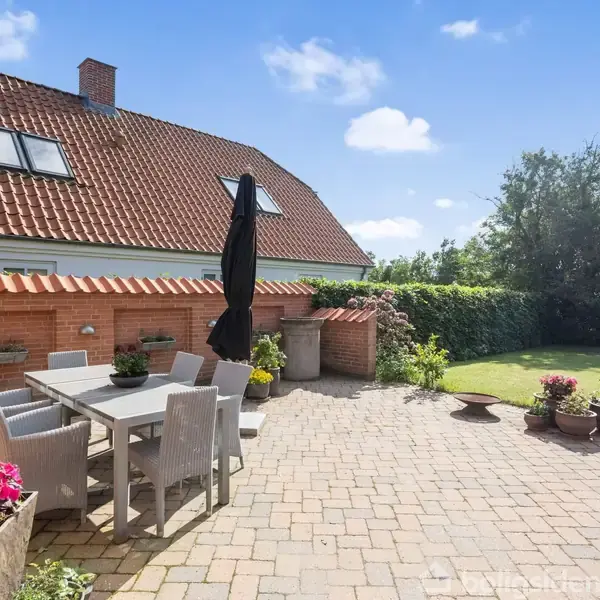 En terrasse med møbler i et grønt haveområde. Der er en grå bord- og stoleopsætning under en sort parasol. Huset i baggrunden har rødt tegltag.