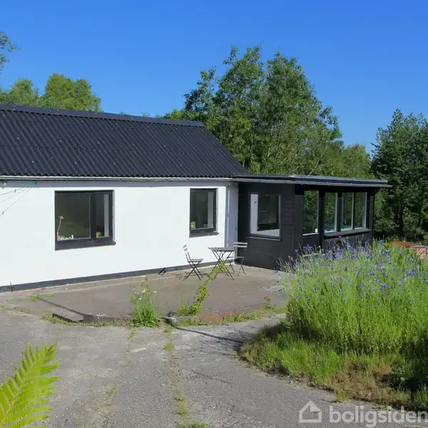 En hvid, ensidet hus med sorte tagsten og flere vinduer står i et grønt, træfyldt landskab. Foran huset er en terrasse med et lille bord og to stole.