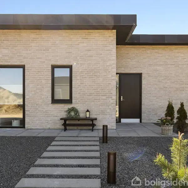 Moderne hus med fladt tag og lys murstensfacade. Foran huset er der en flisebelagt gangsti på småsten. En bænk og planter dekorerer indgangspartiet i sollys.