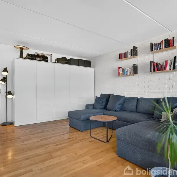 En stor blå sofa står i hjørnet af en stue med hvidmalede murstensvægge. Et rundt sofabord er foran sofaen. Der er bogreoler med bøger og lamper i rummet.