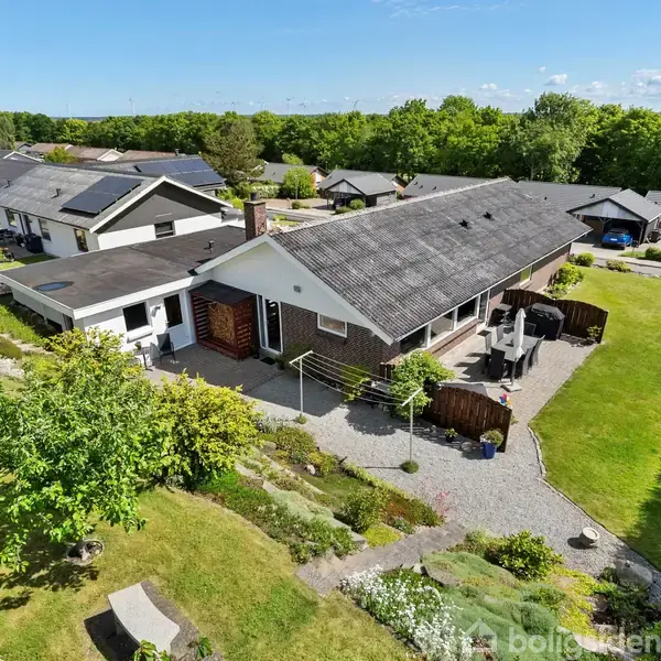 Et stort parcelhus med skråt tag står omgivet af en velplejet have. Huset ligger i et boligområde med flere lignende huse og grønne omgivelser.