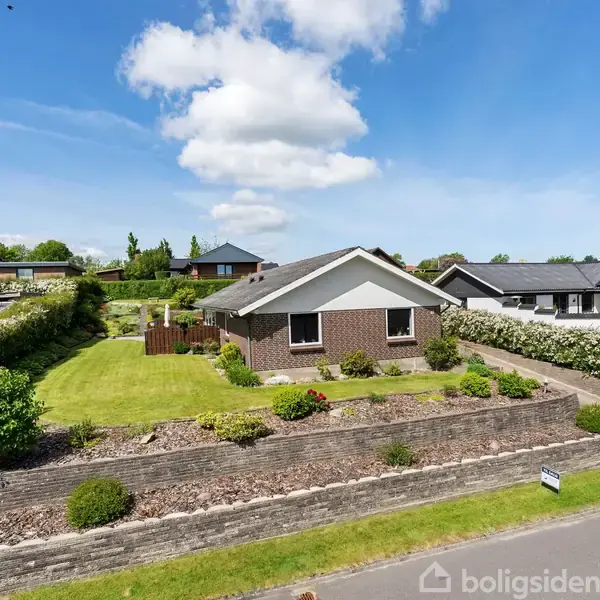 Et murstenshus med hvidt tag står stille i en velplejet have omgivet af buske. Huset ligger i et boligområde med flere lignende hjem i baggrunden under en blå himmel.