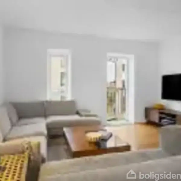 En stue med en stor, grå hjørnesofa står foran et sofabord. En fladskærms-TV hænger på væggen til højre. Gennem vinduerne ses en balkon.