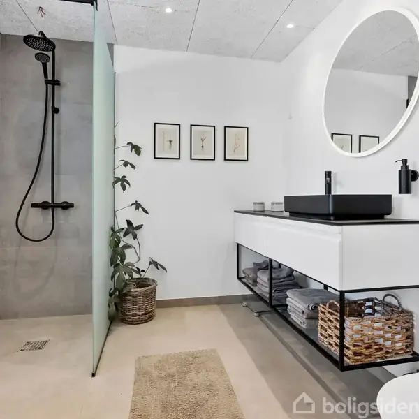 Et moderne badeværelse med en bruseniche til venstre. En sort, rund spejl hænger over en hvid vask på et åbent skab med kurve. Enkel udsmykning med planter og billeder.