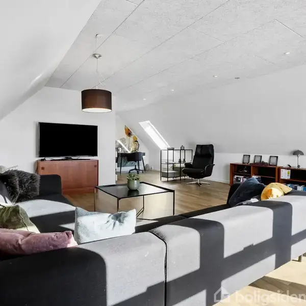 En stor, lys stue med en grå sofa, sofabord og en fladskærms-tv. Rummet har skråvægge, trægulv og flere hylder med bøger i hjørnet.