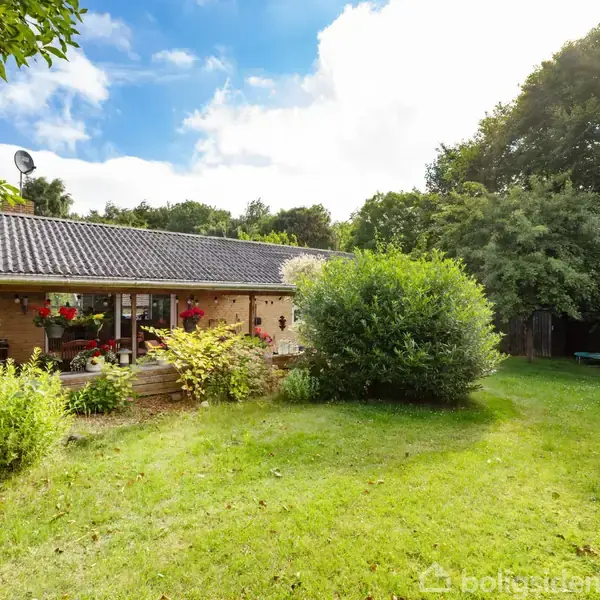 Et hyggeligt hus med gråt tag står omgivet af en frodig have med græs, buske og træer. Huset har en terrasse med blomster og en trædør til venstre.