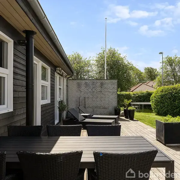 Sort havebord med kurvestole står på en træterrasse ved siden af et mørkt træhus. Terrasse omgivet af grøn have, hæk og få buske. Himmel let skyet.