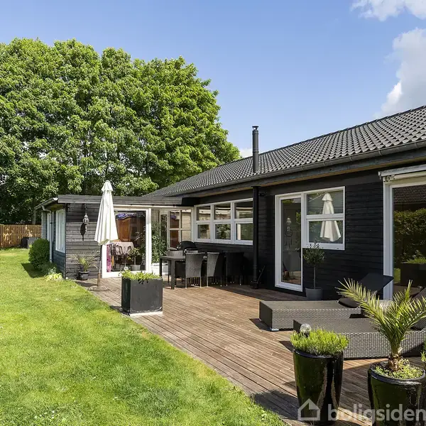 Et sortmalet hus med store vinduer støder op til en terrasse. Terrassen har udendørsmøbler og plantekasser. I baggrunden er der en grøn græsplæne og frodigt løv.