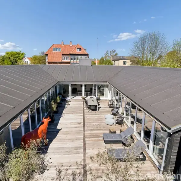 Bygning med mørkt tag omkranser en træterrasse. En orange dekoration ses til venstre. Møbler og planter pryder terrassen. Baggrund viser himmel og omkringliggende huse i grønne omgivelser.