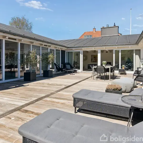 Orange plastikko står stille på træterrasse omgivet af moderne havemøbler og planter i krukker. En række af glasdøre åbner ind til et hus i baggrunden.