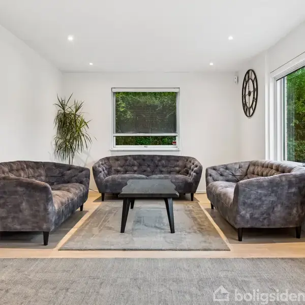 Tre grå sofaer er arrangeret omkring et sort rektangulært sofabord i en lys stue. Et vindue med grøn udsigt og en dekorativ plante pryder rummet.